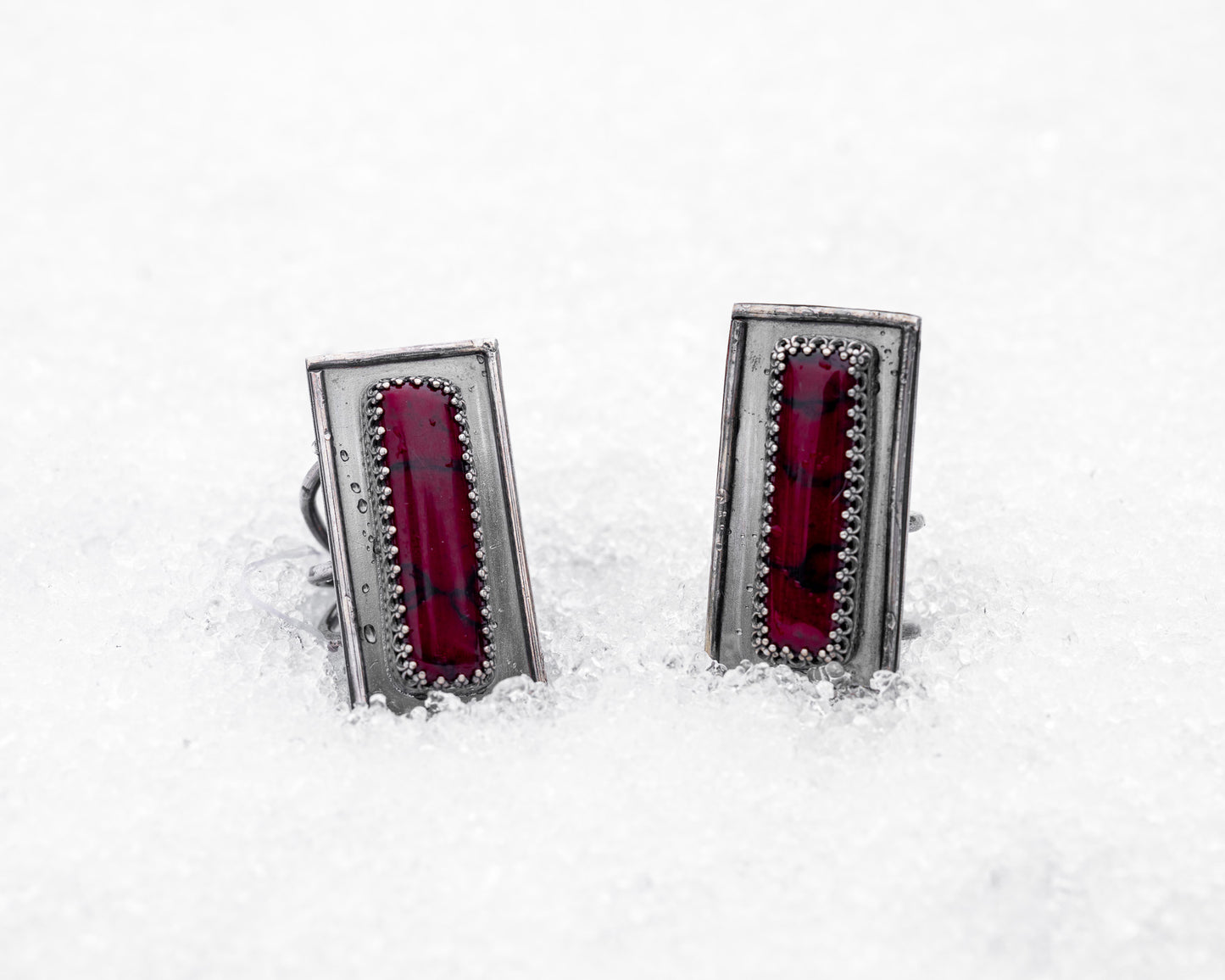 Close-up of a sterling silver ring with an elongated rectangular design set with a deep red bloody bison jasper, nestled upright in textured white snow. The ring has detailed wirework around the stone and a dark, oxidized finish, evoking an ancient, blood-bound artifact.
