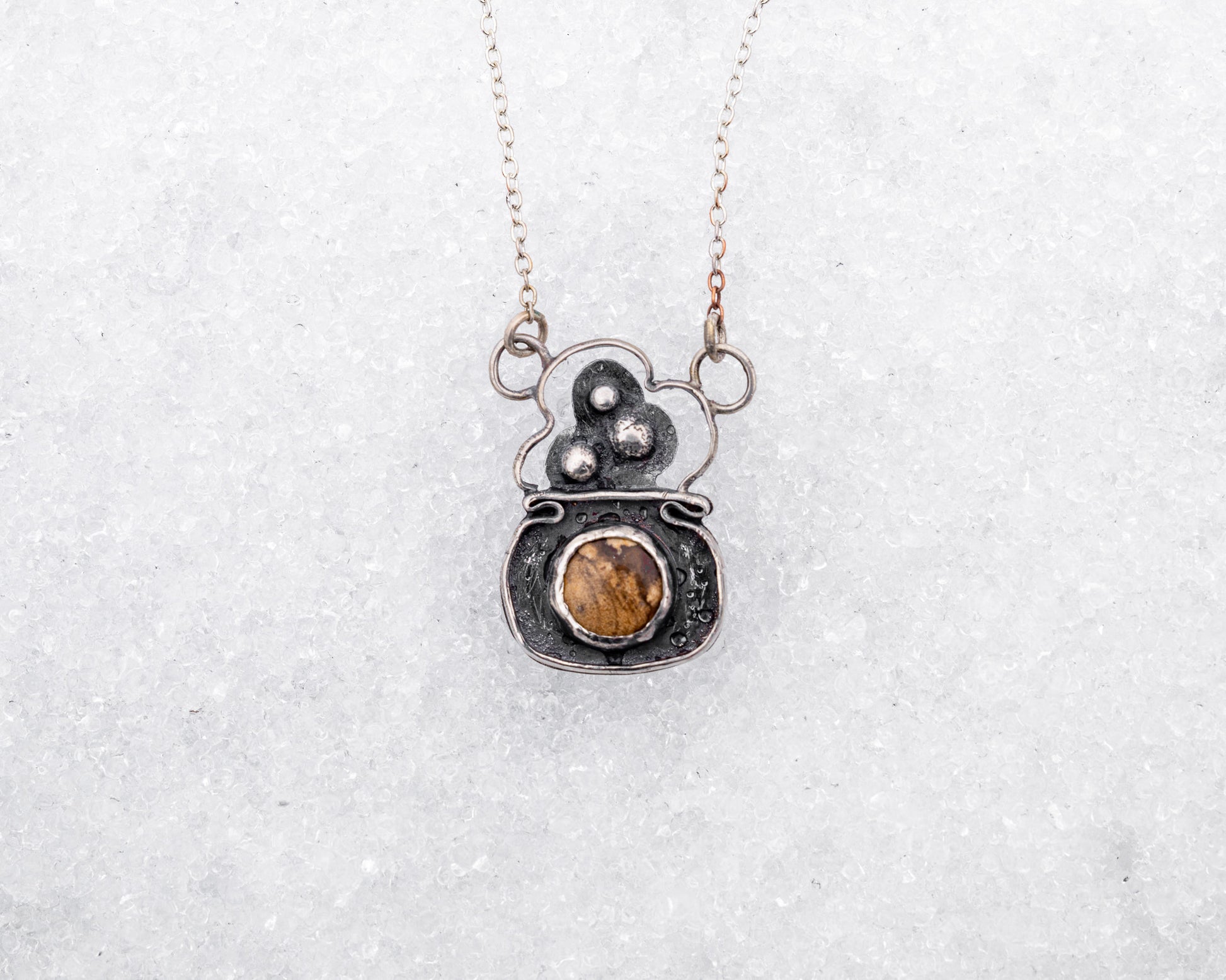 Handcrafted sterling silver necklace with a rounded pendant featuring three raised silver bubbles above a bezel-set garden jasper cabochon. Styled on snow, the pendant resembles a bubbling cauldron used in magical rituals.
